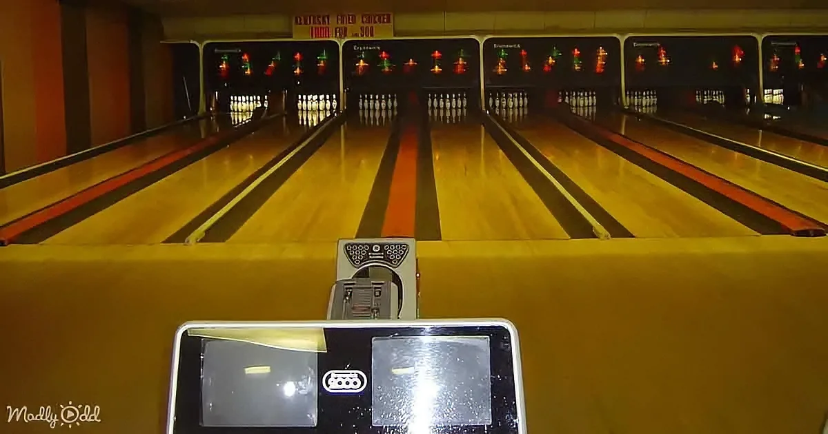 A bowling alley lane with pins set up, a ball return machine in the foreground, and a sign above the lanes.