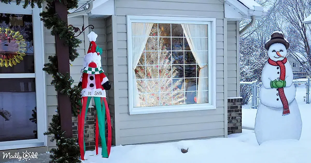 A festive snowman decoration stands in front of a house with a decorated window and a tall, colorful snowman figure nearby.