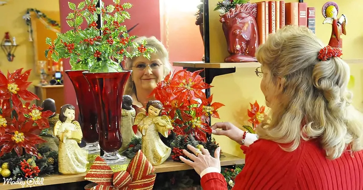 A woman in a red sweater arranges Christmas decorations, including poinsettias and angel figurines, on a shelf.