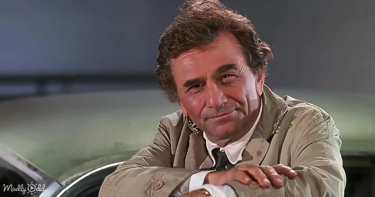 A man in a beige jacket leans on a car, smiling with a hand on his chest.