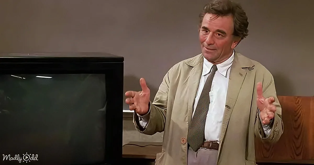 A man in a beige suit gestures with his hands while standing next to a black television.