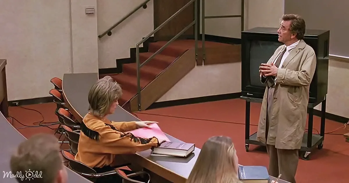 A man in a tan coat stands in a classroom, gesturing, while a woman sits at a desk, holding a book.