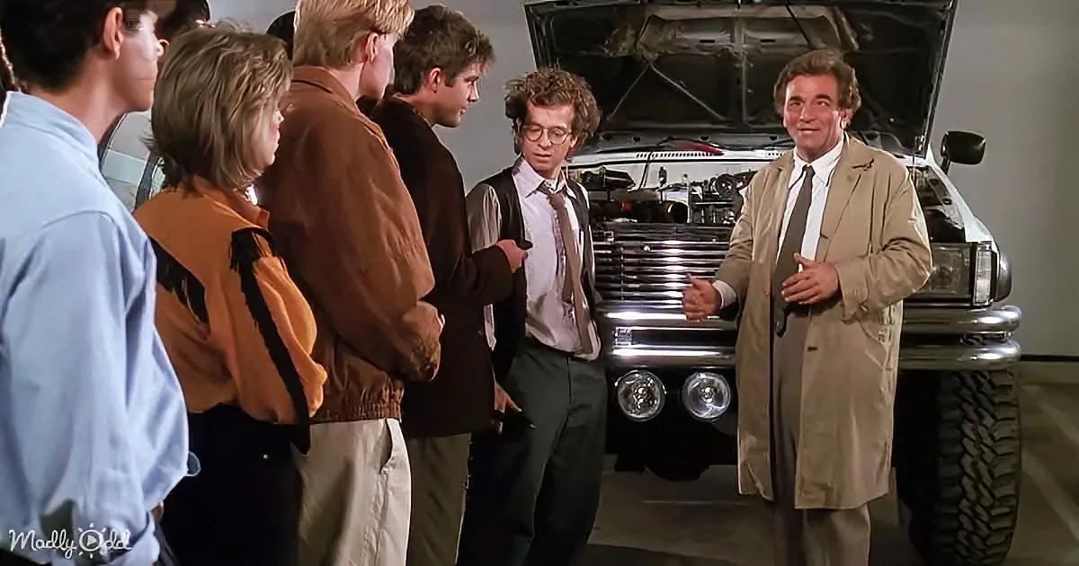 A man in a trench coat stands by a car with an open hood, talking to a group of people.