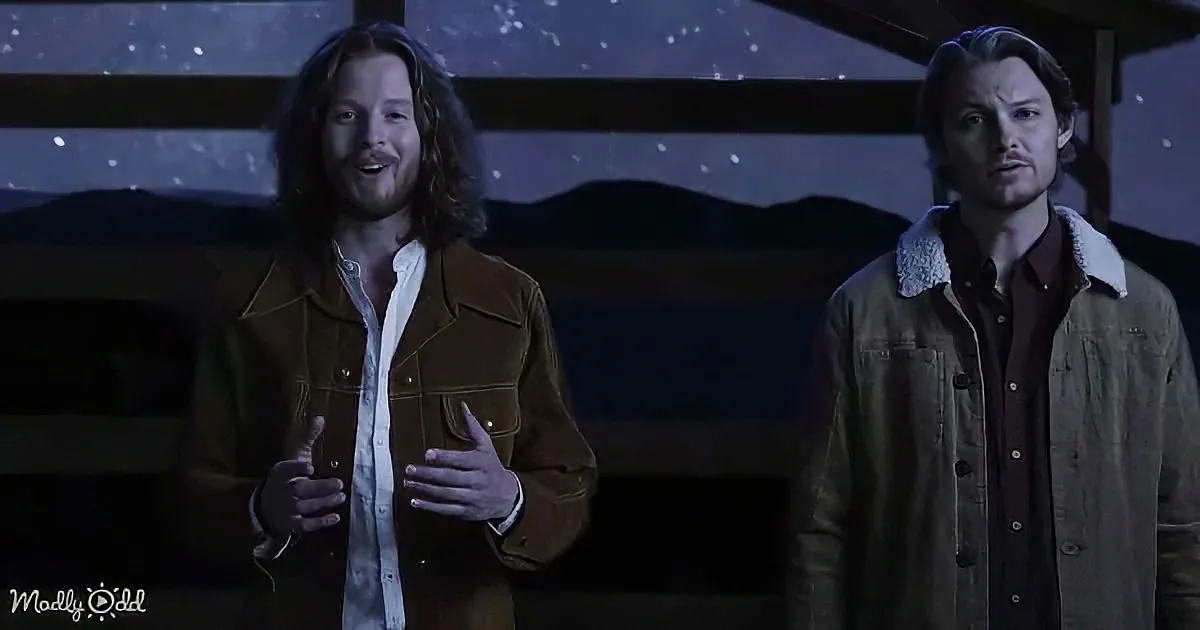 A man with long hair and a beard gestures while another man stands beside him, both in jackets, against a starry night...