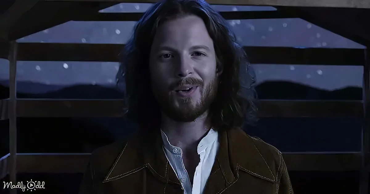 A man with long hair and a beard, wearing a brown jacket and white shirt, stands in front of a window with a starry night...