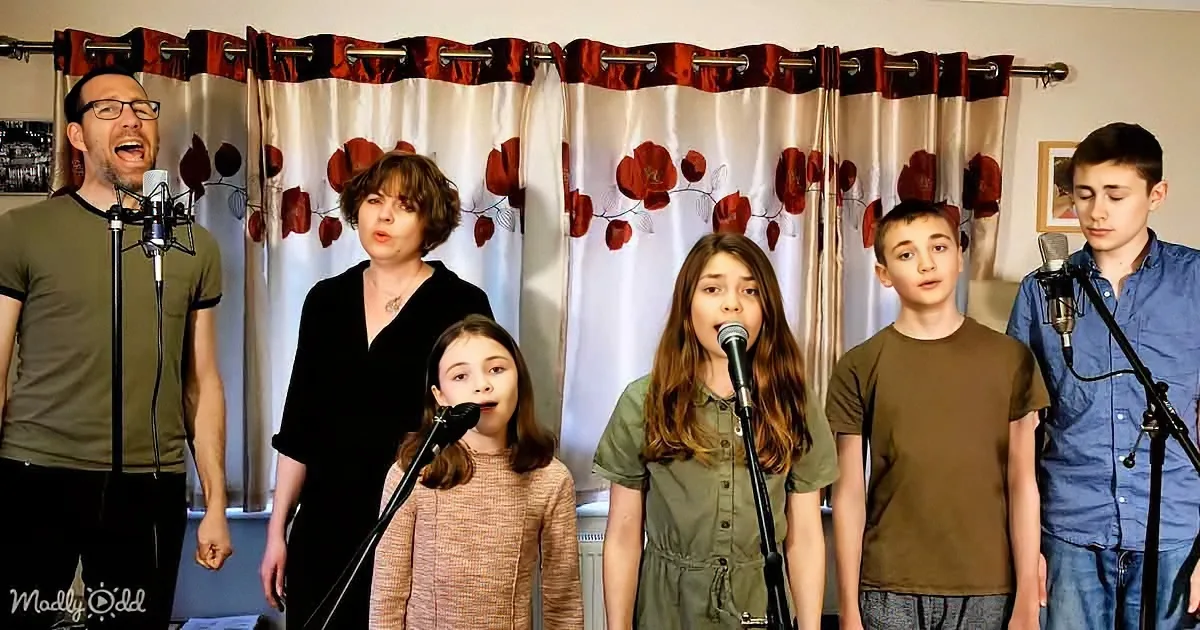 A group of six people, including children, sing into microphones in front of a floral curtain backdrop.