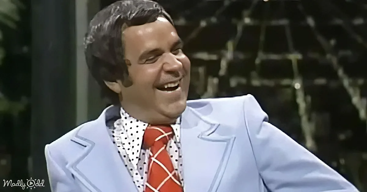 A man in a light blue suit with a red tie and white polka dot shirt is smiling.