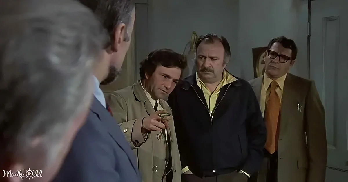 A man in a trench coat holds a gun, facing another man in a suit, with two others in suits and ties observing.