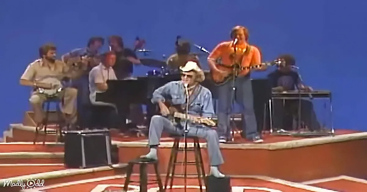 A man in a cowboy hat sits on a stool, playing guitar on a stage with a band behind him.