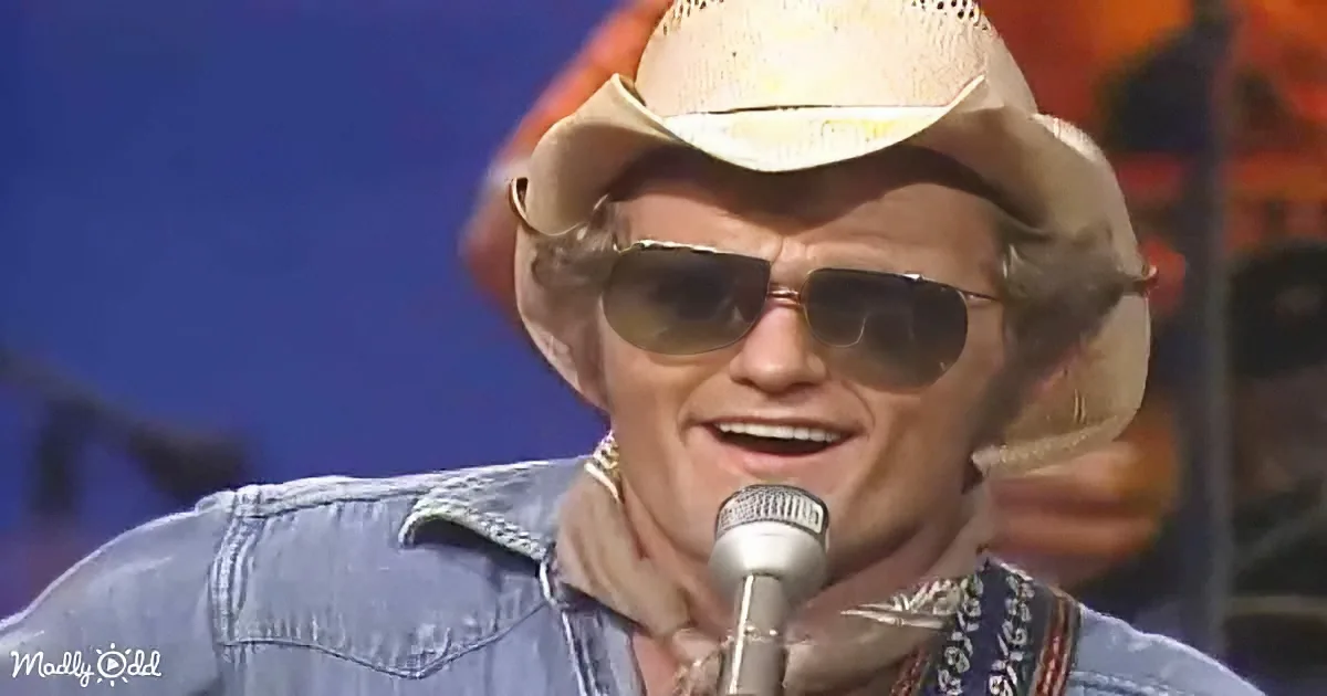 A man wearing a straw hat and sunglasses sings into a microphone, smiling.