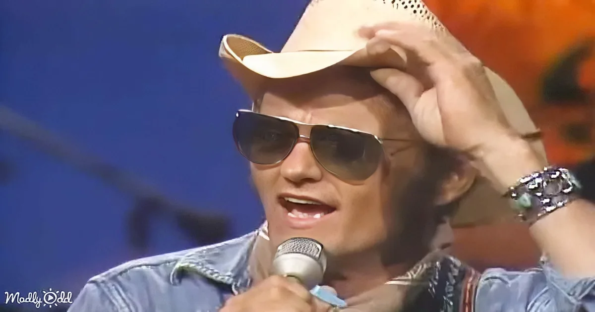 A person wearing a cowboy hat and sunglasses sings into a microphone, adjusting their hat with one hand.