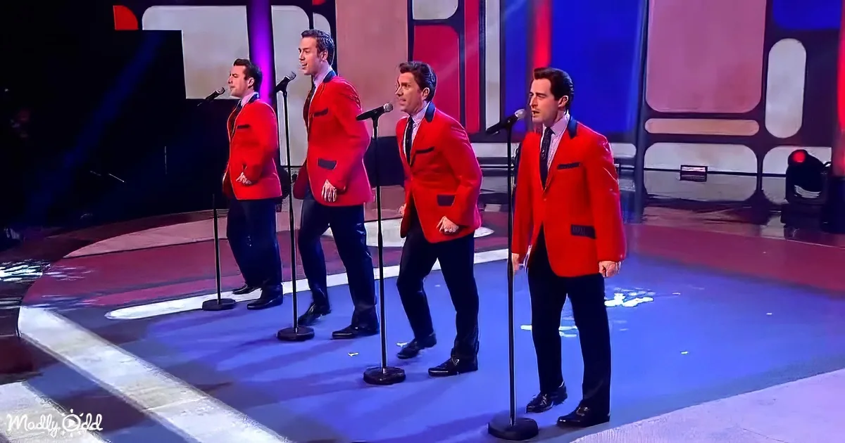 Four men in red suits sing on stage with microphones, wearing black pants and ties.