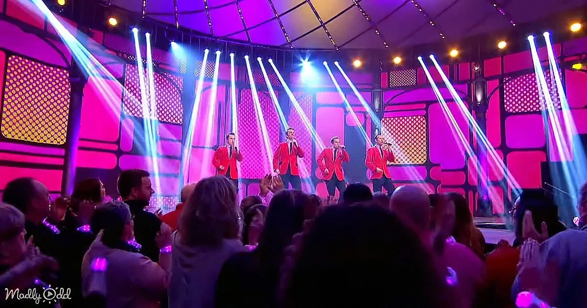 Four men in red suits sing on a stage with bright blue and purple lights, surrounded by an enthusiastic audience.