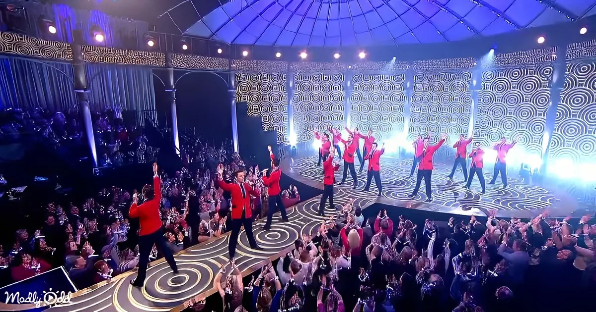 A group in red jackets performs on a stage with a spiral pattern, surrounded by an enthusiastic crowd.