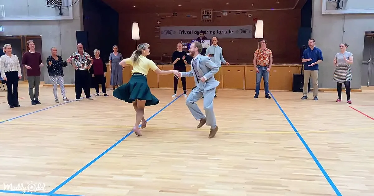 A couple dances in a gymnasium, surrounded by onlookers, with a banner reading "Trivsel og oplevelser for alle" in the...