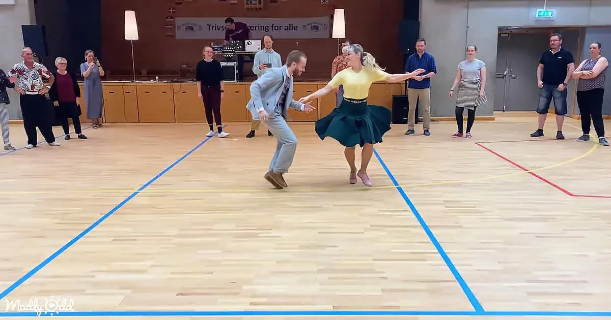 A man and woman dance in a gymnasium, surrounded by onlookers, with a banner in the background.