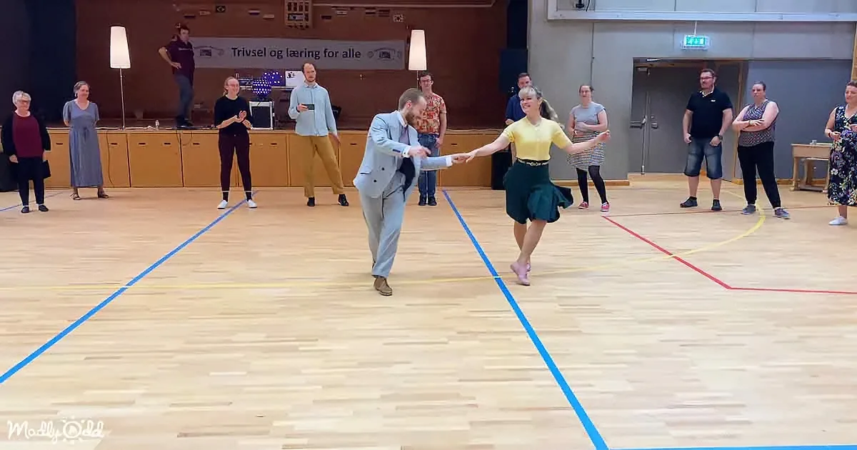 A man and woman dance in a gymnasium, surrounded by onlookers, with a banner in the background.
