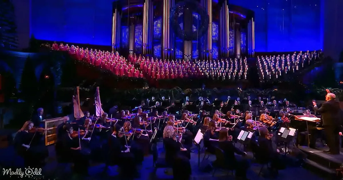 A grand orchestra and choir perform in a dimly lit, ornate concert hall with blue lighting.