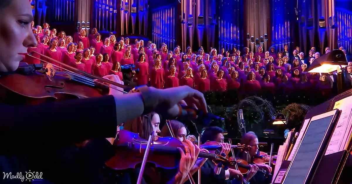 A violinist plays in front of a choir and orchestra in a grand hall.