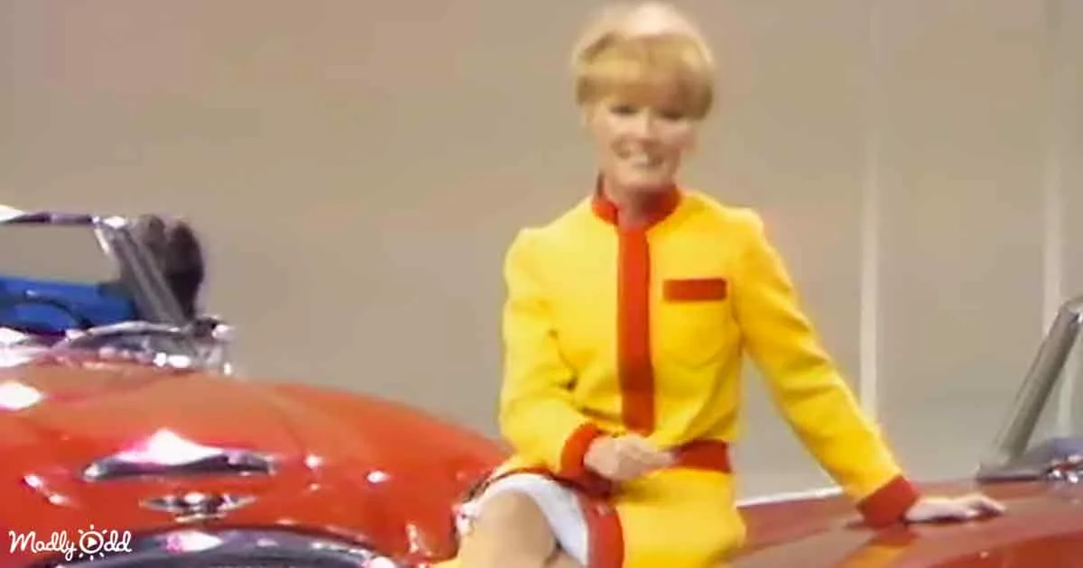 A person in a yellow suit sits on a red car, smiling, with a blurred background.