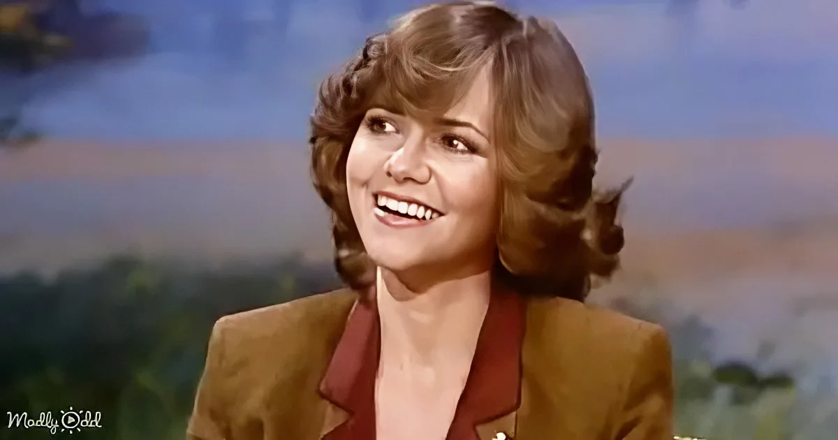 A smiling woman with curly hair, wearing a brown blazer and red shirt, against a scenic backdrop.