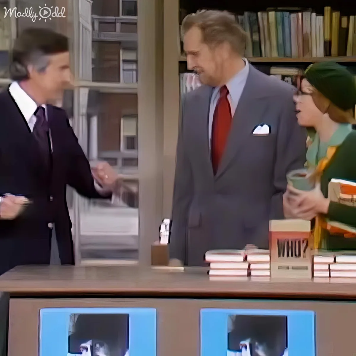 A man in a suit and tie is shaking hands with another man in a suit, with books and a "Who?" book visible on a desk.