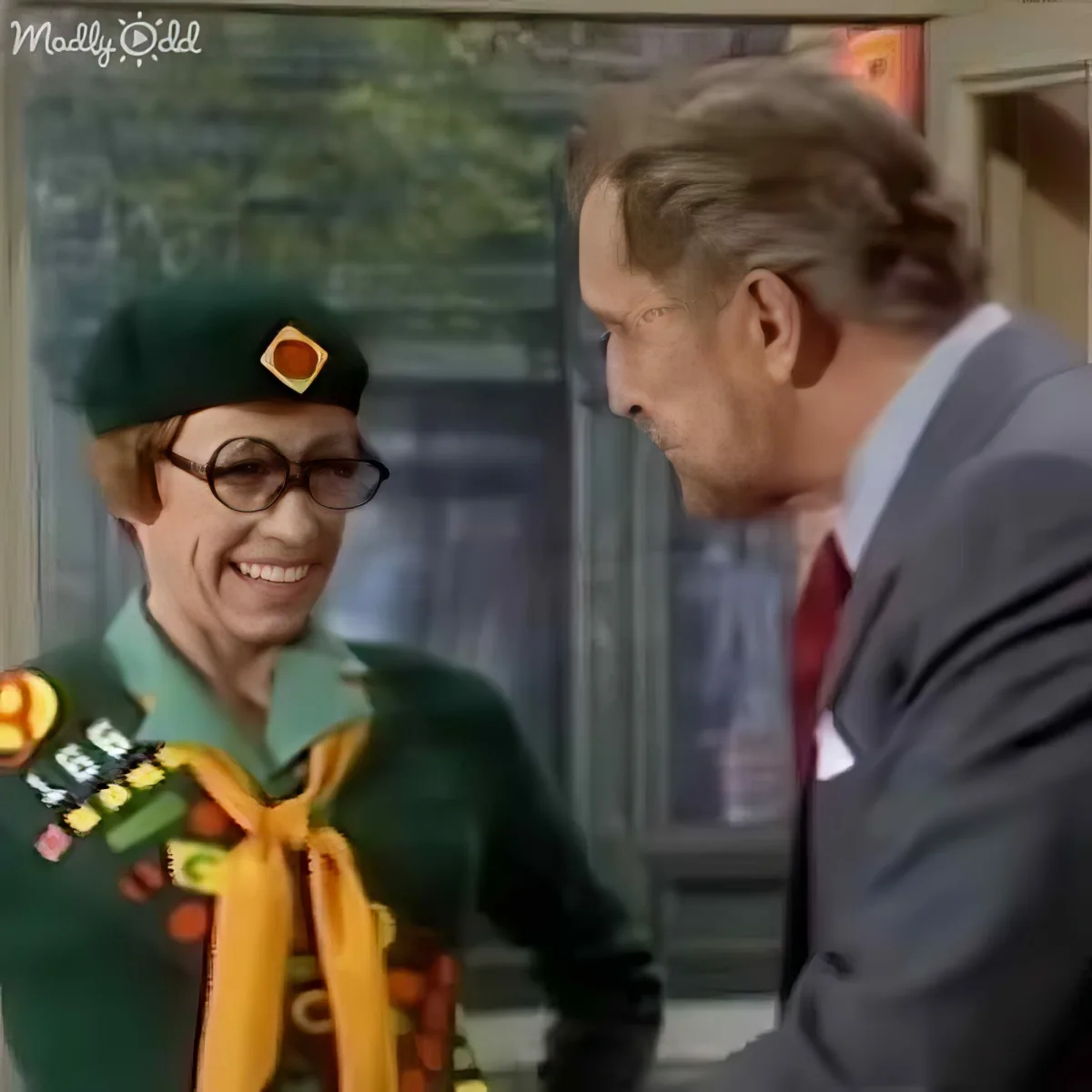 A person in a green scout uniform with medals and a hat is smiling at another person in a suit.