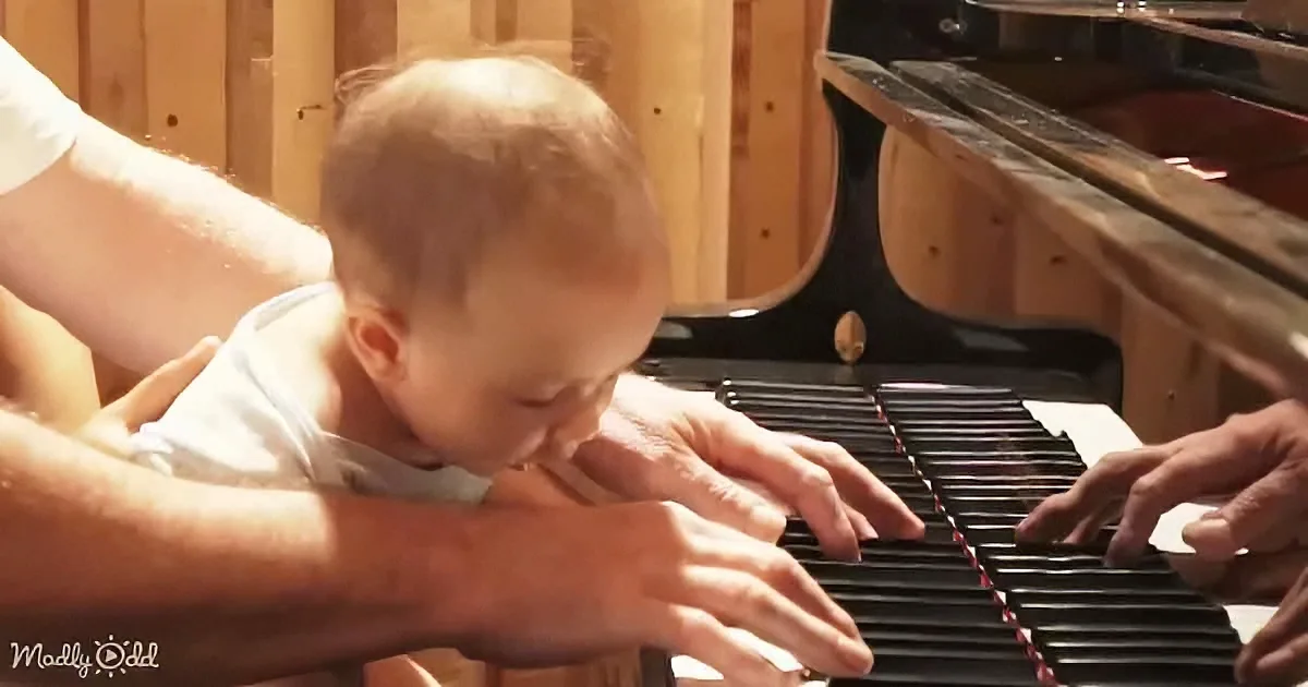 A baby leans over a piano keyboard, hands on keys, with an adult's hands nearby.