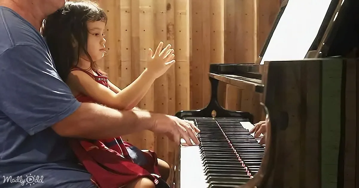 A child sits on an adult's lap, both playing a piano together in a sunlit room.