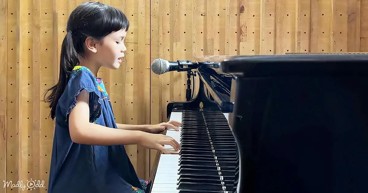 A young girl in a blue dress plays a grand piano with a microphone in front.