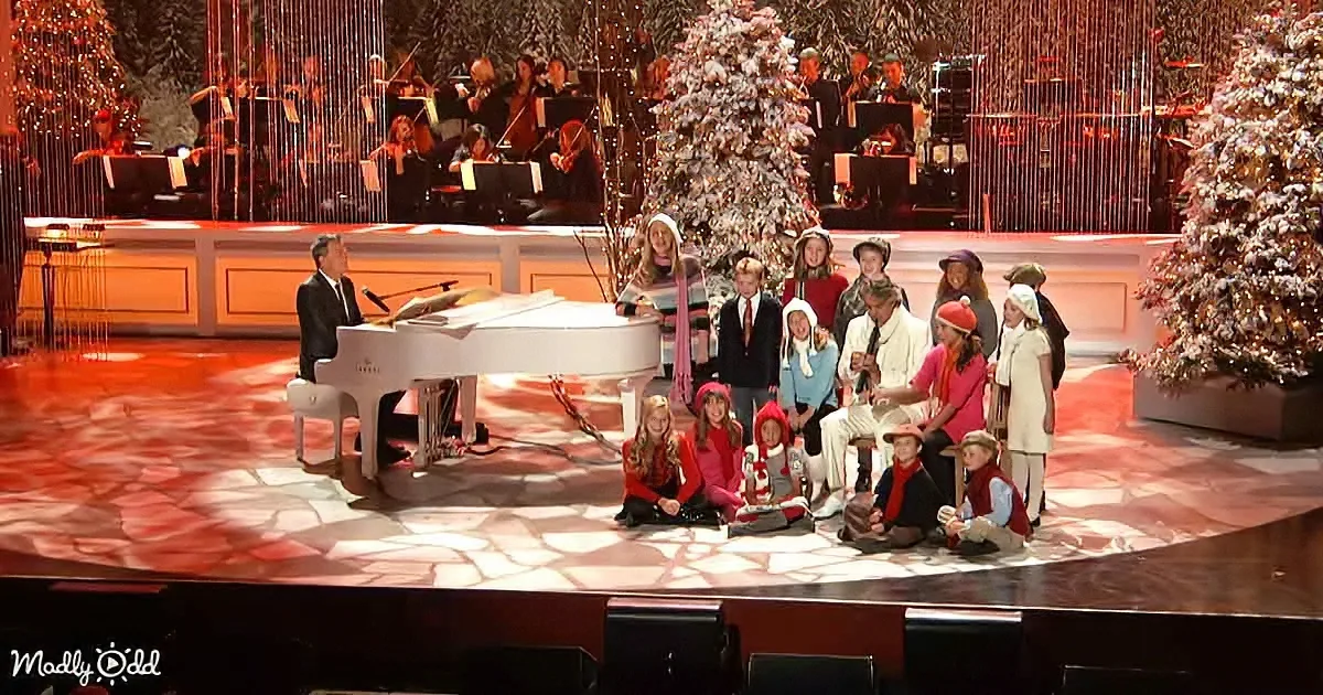 A man plays piano on stage with children in festive attire, surrounded by Christmas trees and an orchestra.