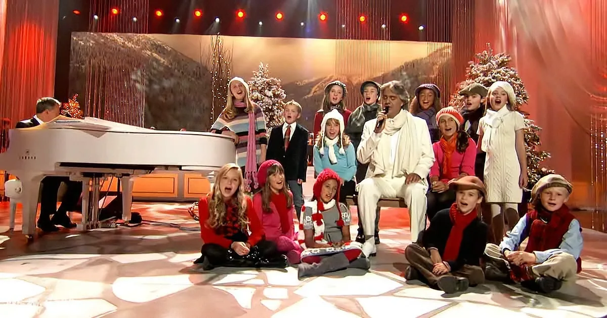 A man in white sings into a microphone, surrounded by children in festive attire, with a piano and Christmas trees in the...