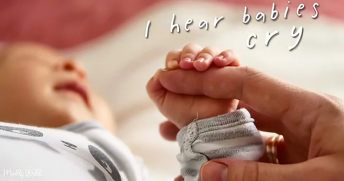 A baby's hand is being held by an adult's hand, with the baby lying down and wearing a white outfit.