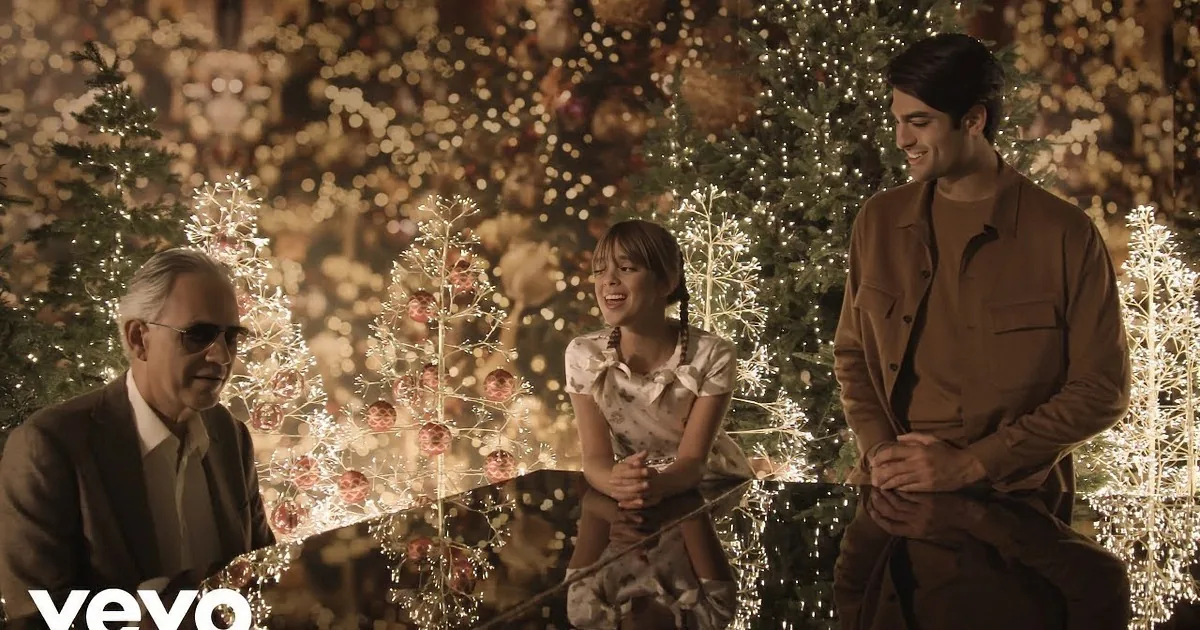 A man plays piano amidst a festive Christmas setting with two others, surrounded by illuminated trees and ornaments.