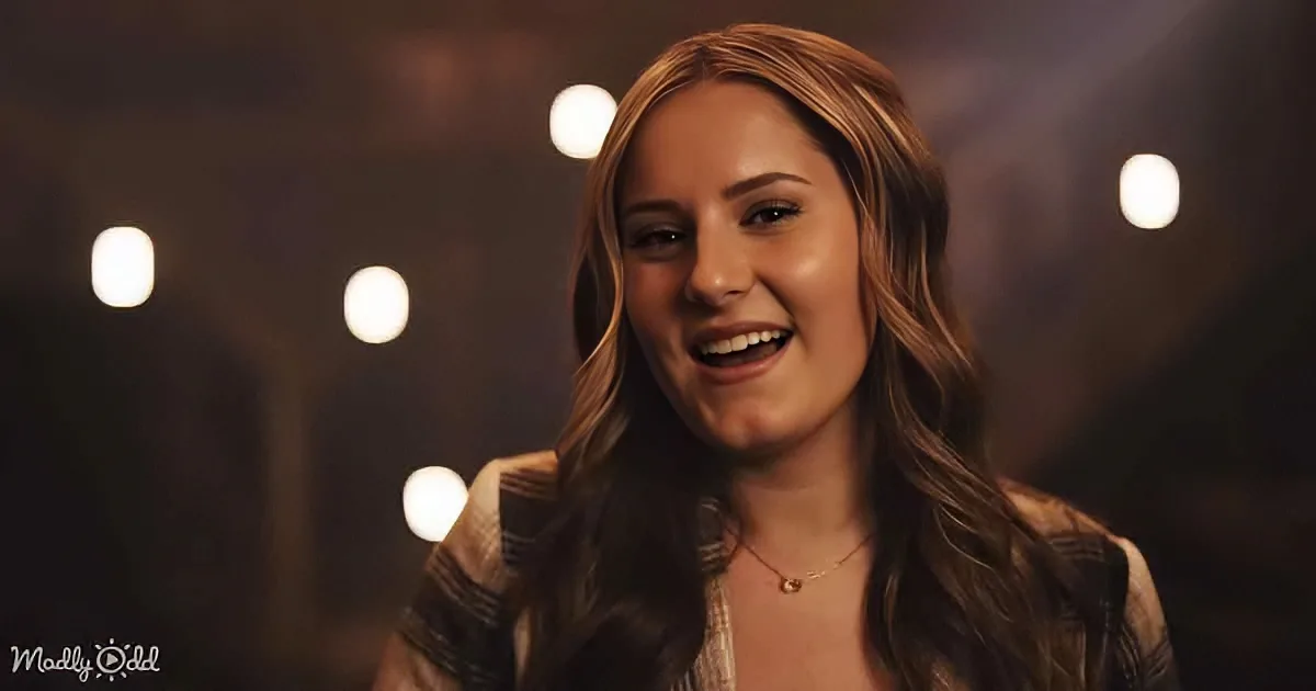 A smiling woman with long brown hair, wearing a striped shirt and a necklace, stands in front of blurred lights.
