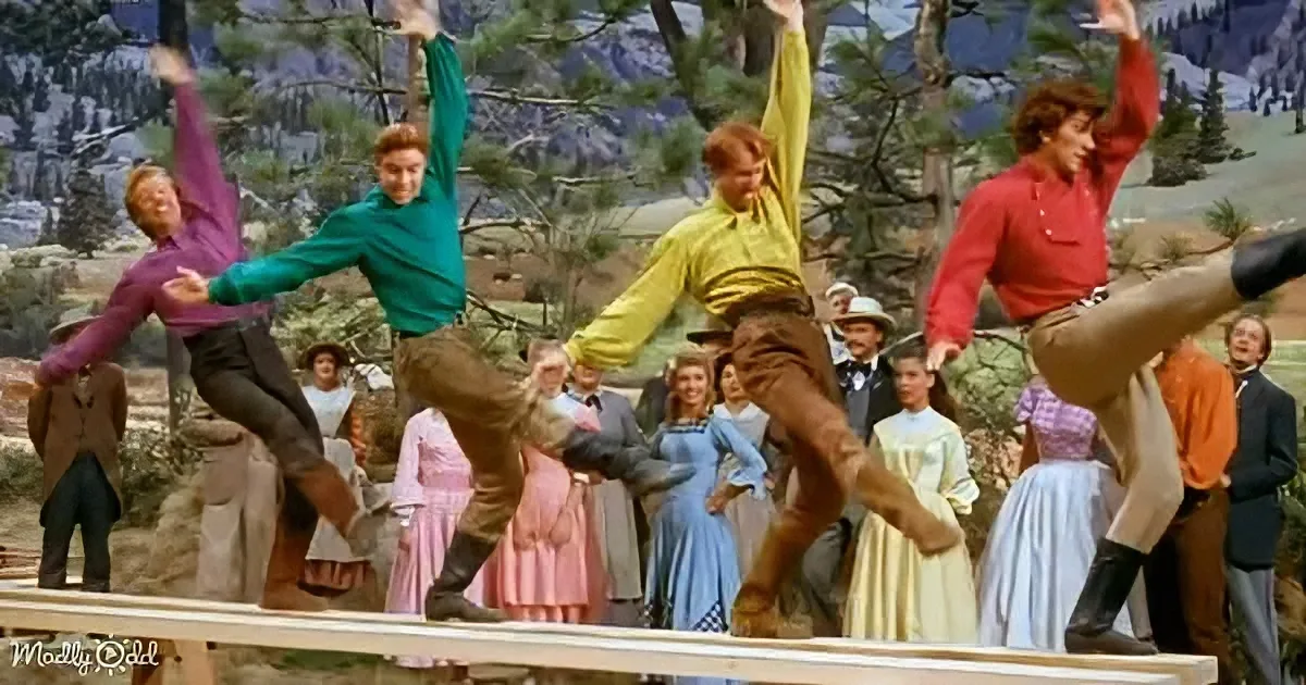 Men in colorful shirts perform a synchronized dance on a wooden beam in front of a scenic backdrop.
