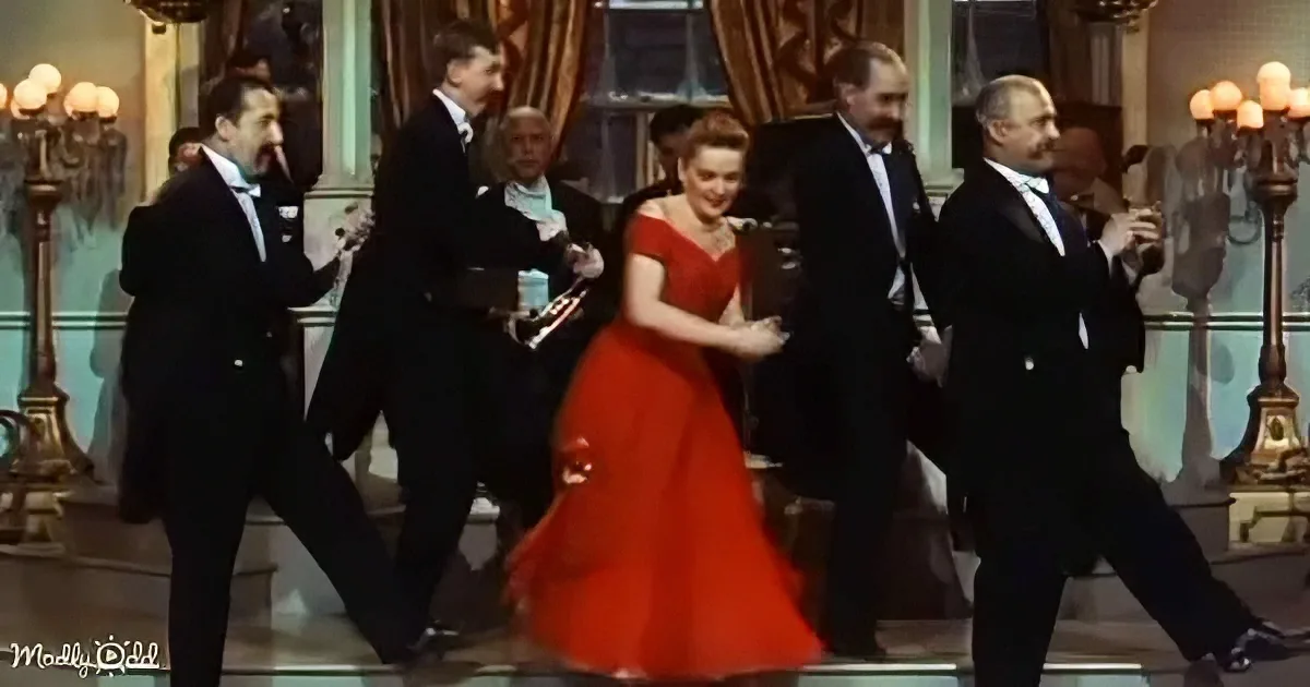 A woman in a red dress dances surrounded by men in tuxedos in a grand, ornate room.