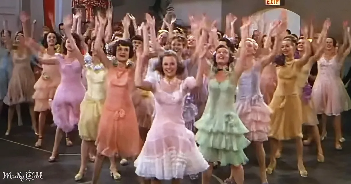A woman in a white dress leads a group of women in colorful dresses in a synchronized dance.