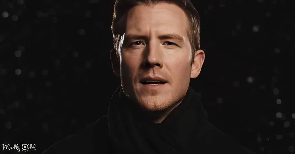 A man with short brown hair, wearing a dark jacket, stands against a dark background with small white dots.