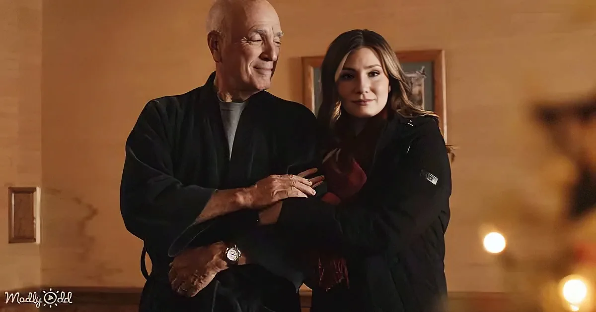 An older man in a black robe holds a woman's hand, both smiling softly in a warmly lit room.