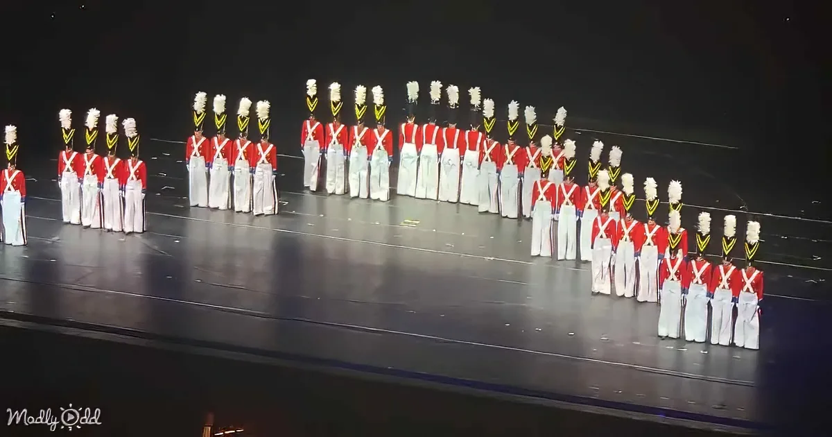 A group of performers in red and white uniforms with plumed hats stand in formation on a stage.