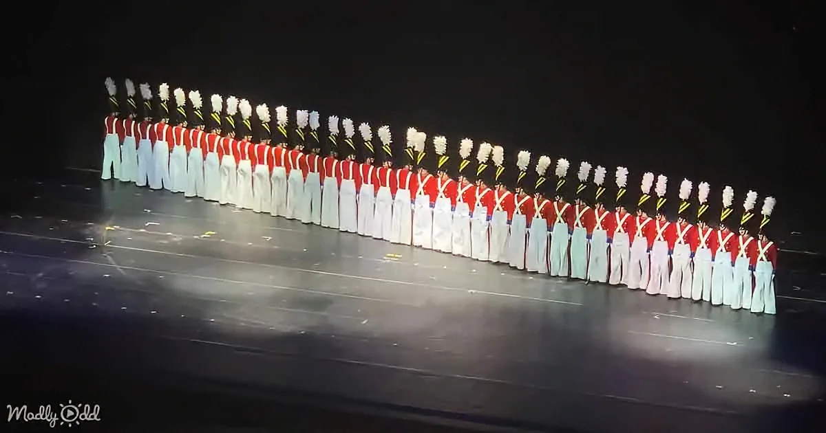 A line of uniformed performers in red and white stands in formation on a dark stage.