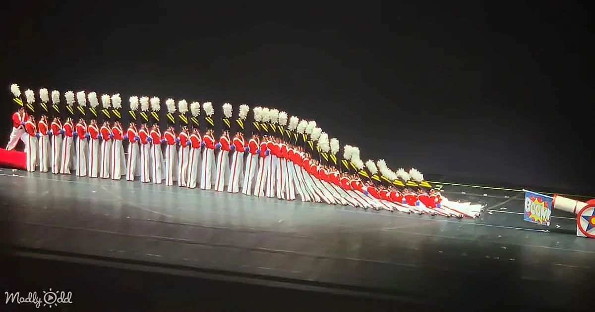 A marching band in red and white uniforms forms a pyramid on a dark stage.