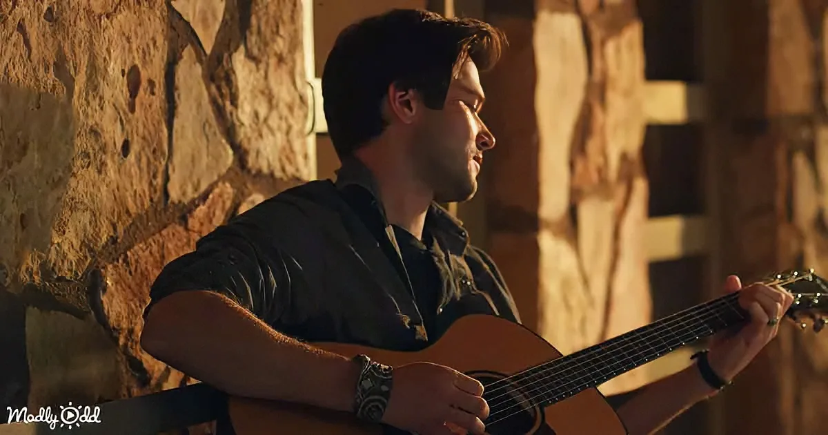 A man in a black shirt plays an acoustic guitar against a stone wall.