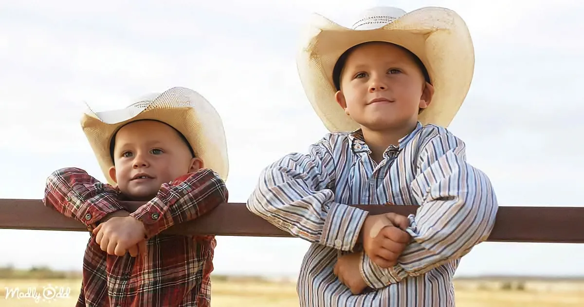 Two young boys in cowboy hats lean on a fence, one in plaid, the other in stripes, against a clear sky backdrop.