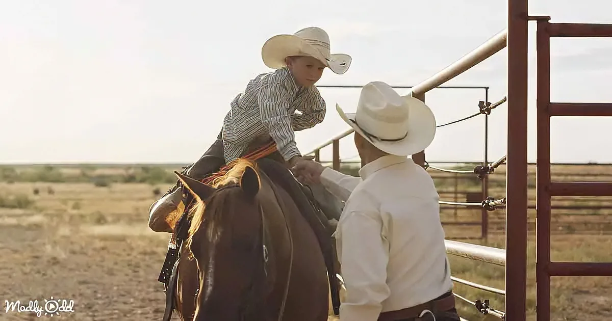 A young boy in a cowboy hat is being helped onto a horse by an adult in a corral.