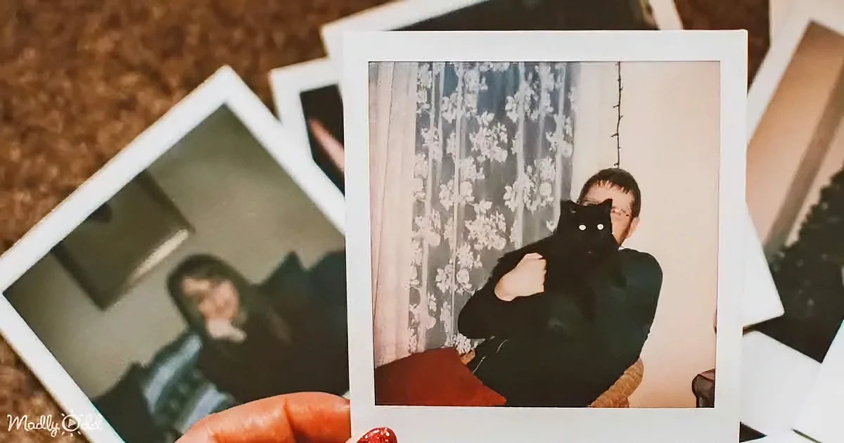 A hand holds a Polaroid photo of a person with a black cat, surrounded by other Polaroids on a textured surface.