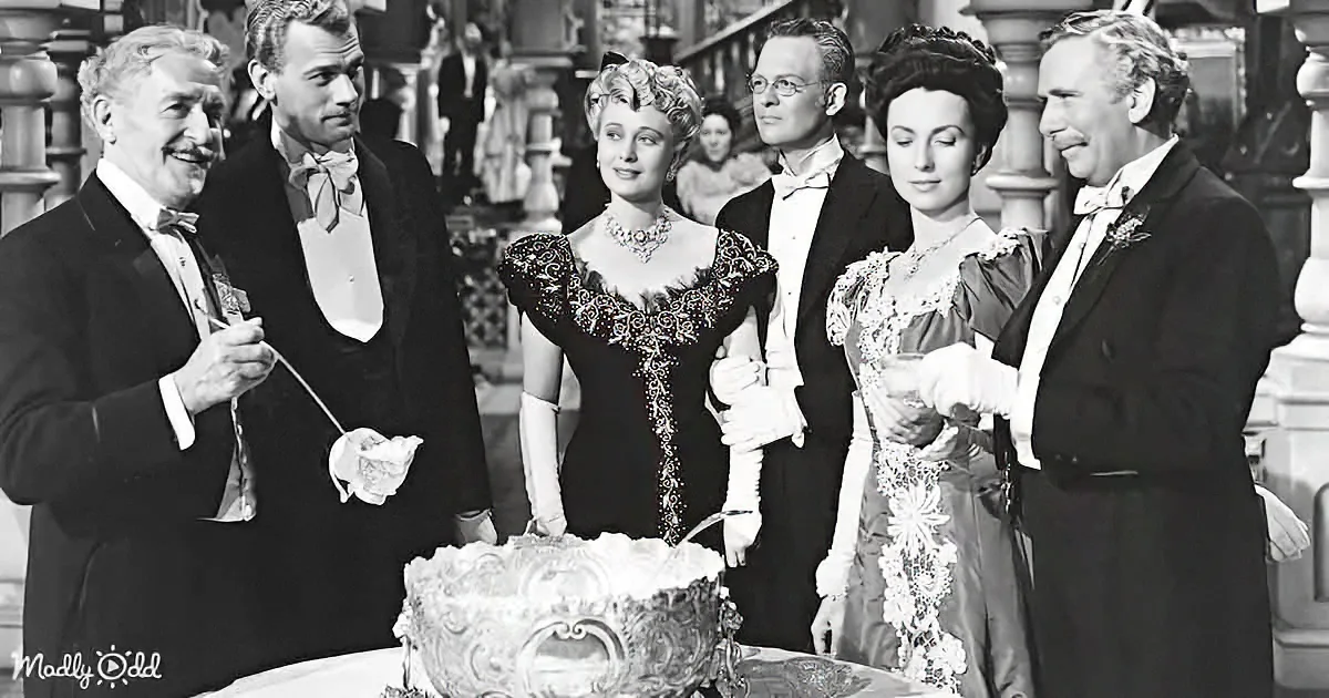 A group of elegantly dressed individuals gather around a large, ornate cake, seemingly preparing to cut it.