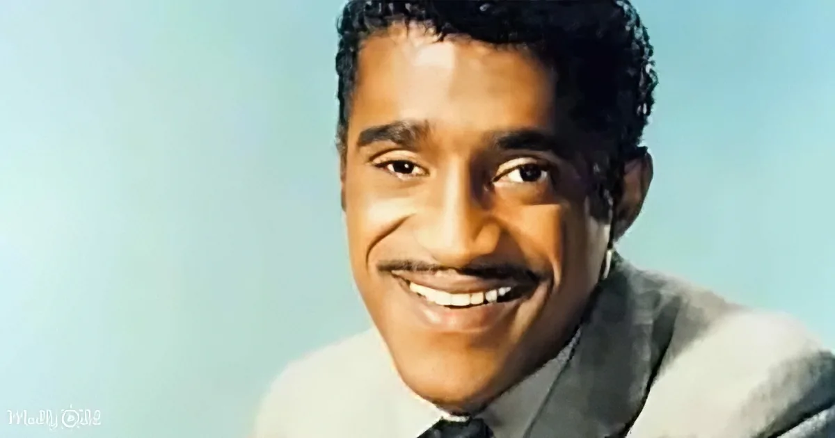 A smiling man with short, curly hair, wearing a light-colored suit and tie, against a light blue background.