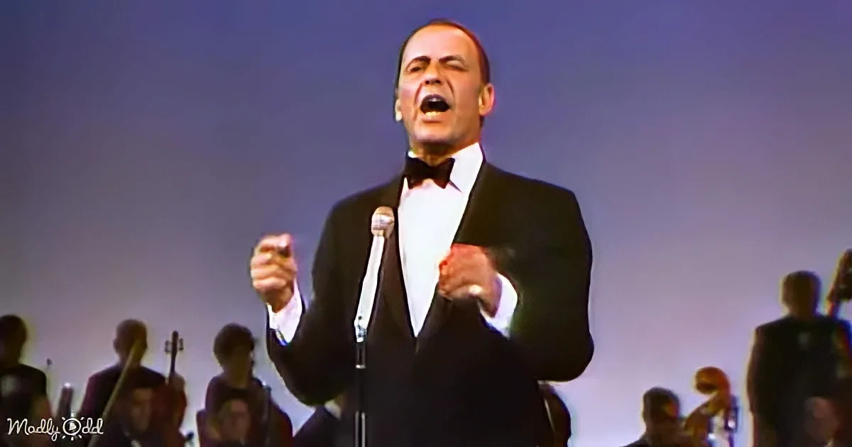 A man in a tuxedo sings passionately on stage with an orchestra in the background.